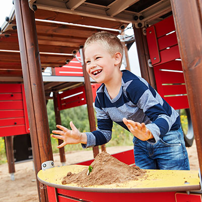 Ein kleiner Junge spielt mit Sand unter einem großen Multispiel Spielanlage. Es gibt eine Theke im Stil eines Spielhauses, in dem die Kinder fantasievoll spielen können.